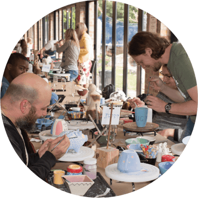 Team members engaging in a creative pottery painting activity at a long table in a bright, open space with large windows, fostering collaboration and a fun, relaxed atmosphere.