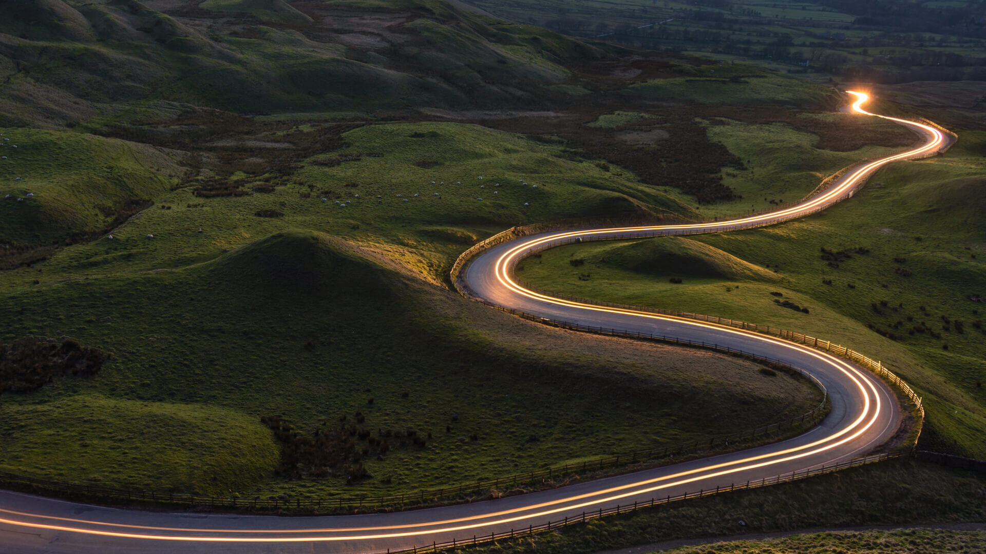 A winding mountain road at dusk or dawn, captured using long exposure photography. The road snakes through green rolling hills and is marked by continuous light trails from vehicle headlights and taillights, creating a glowing S-curve that stretches into the distance. The road is bordered by wooden fencing, and the surrounding landscape features lush green hillsides in low light conditions.