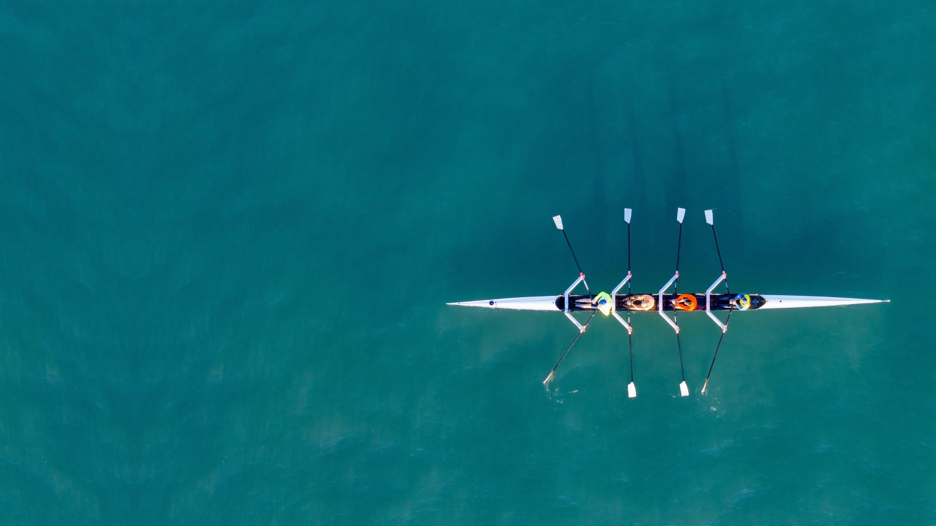 An aerial view of a rowing team in a white racing shell on turquoise water. The image shows four rowers in synchronized position with their oars extended, captured from directly above. The rowers are wearing colorful uniforms that contrast against the white boat and deep turquoise water background.