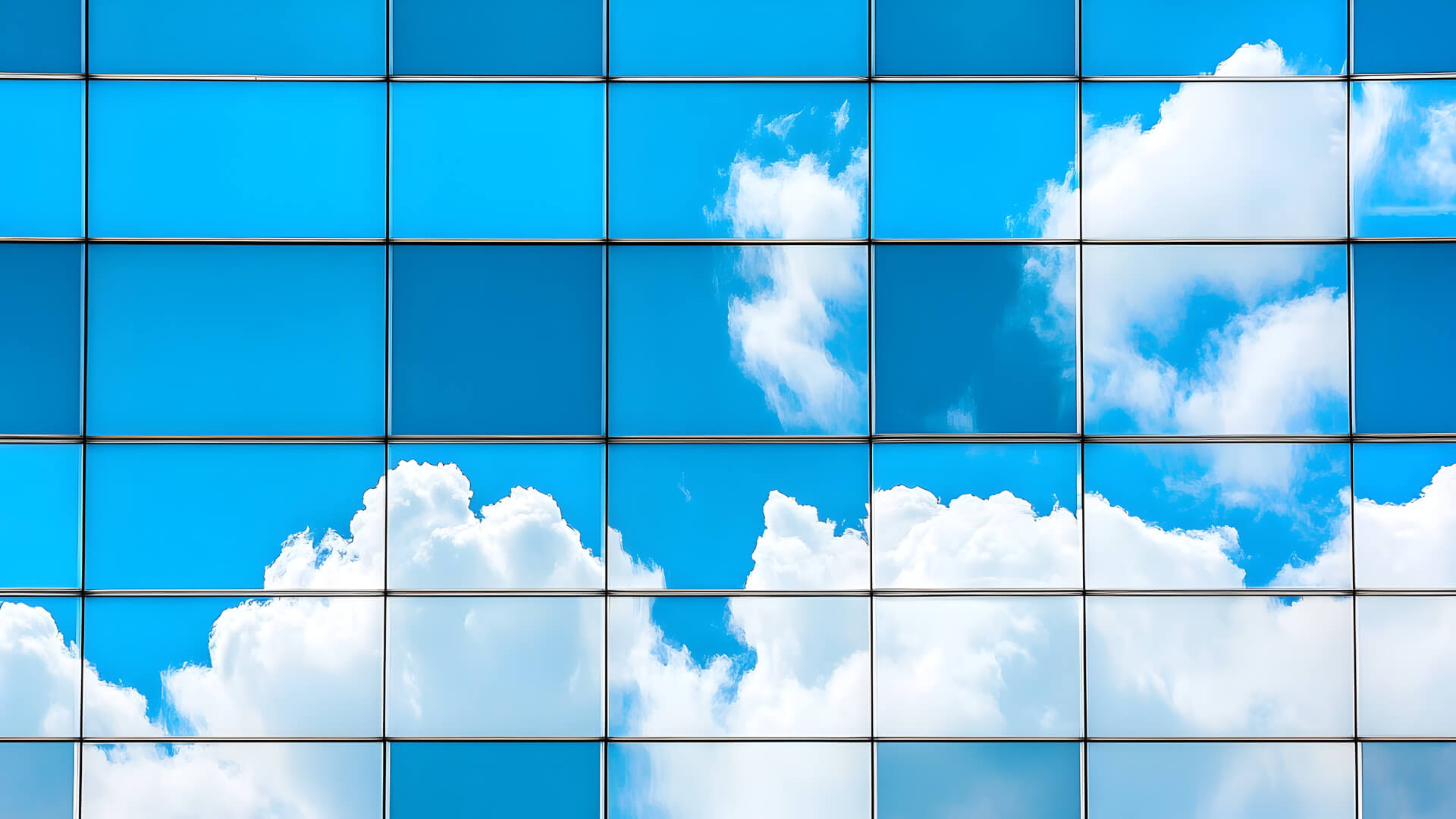 A modern glass building facade reflecting a bright blue sky with white fluffy clouds. The facade is divided into a grid of rectangular glass panels, each mirroring portions of the sky and clouds, creating a geometric pattern of blue and white reflections.