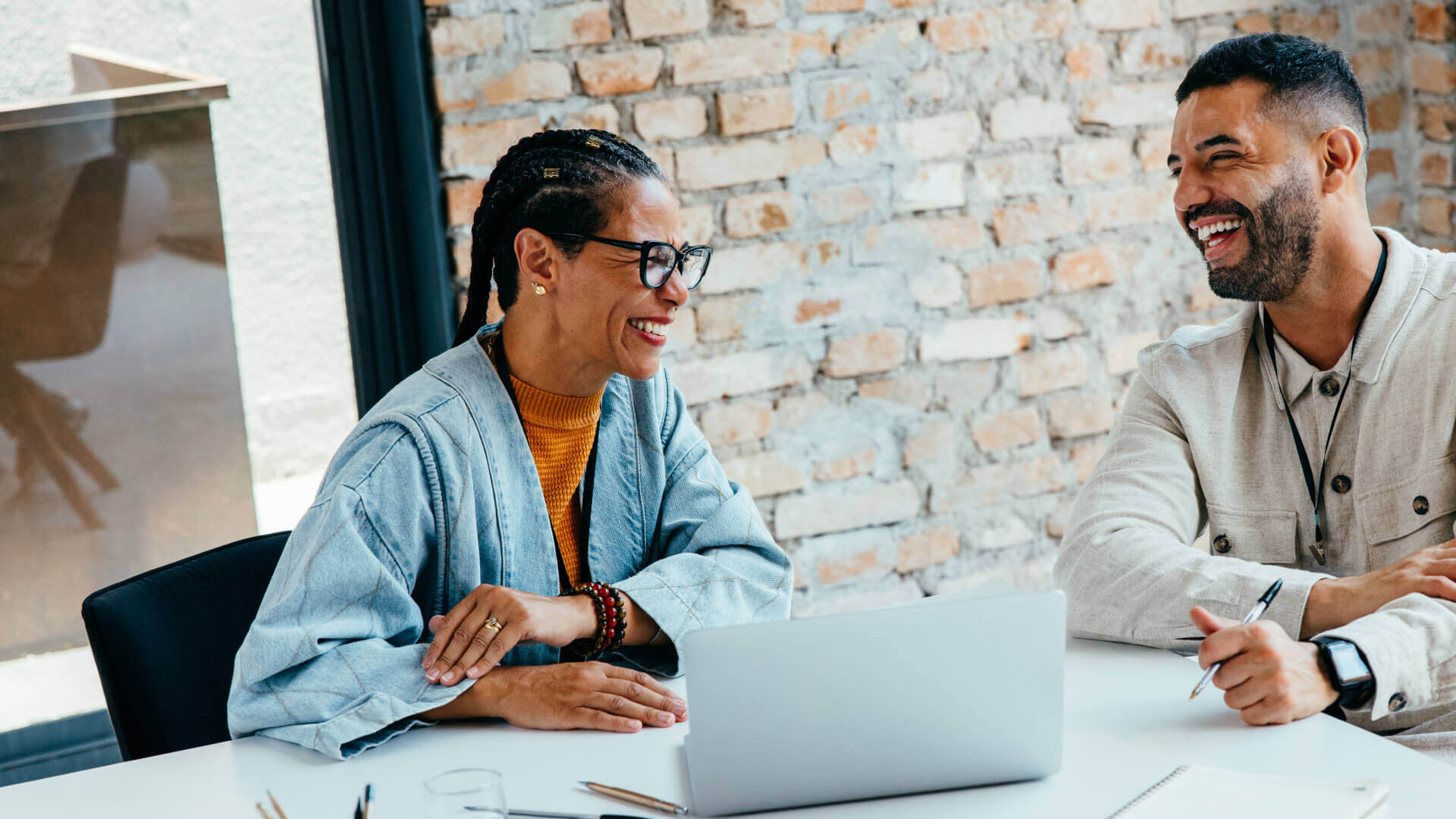 This image shows two people sharing a lighthearted moment during what appears to be a work meeting or collaboration session. A woman wearing glasses, braided hair, an orange sweater, and a light blue denim jacket is seated at a table with a laptop. Across from her is a man in a light-colored button-up jacket, holding a pen. Both are laughing genuinely, suggesting they're having an enjoyable interaction. They're in what looks like a modern office space with exposed brick walls.