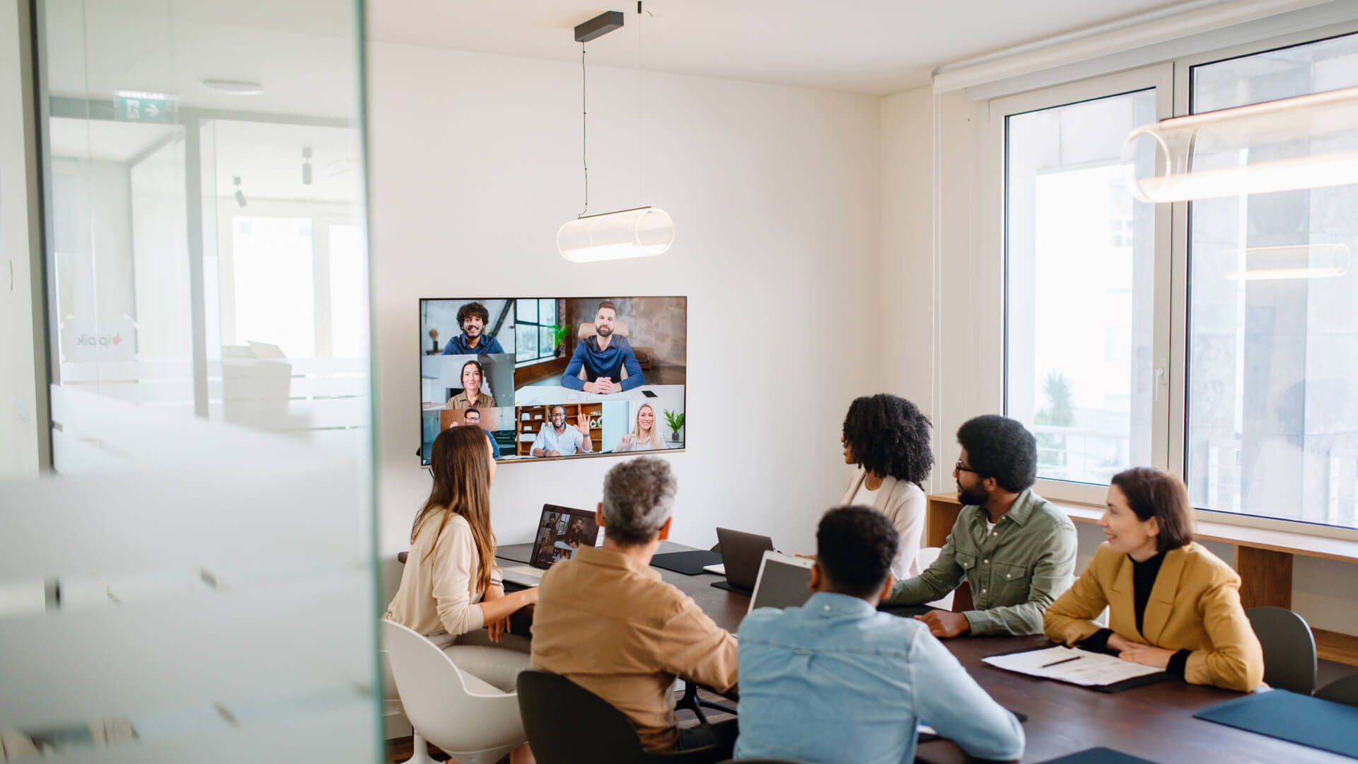 This image shows a hybrid meeting setup in a modern office conference room. Several people are seated around a conference table, viewing a wall-mounted TV screen displaying a video conference call with multiple remote participants in a grid layout. The in-person attendees appear to be diverse, including people wearing various business casual attire in colors like yellow, beige, and green.