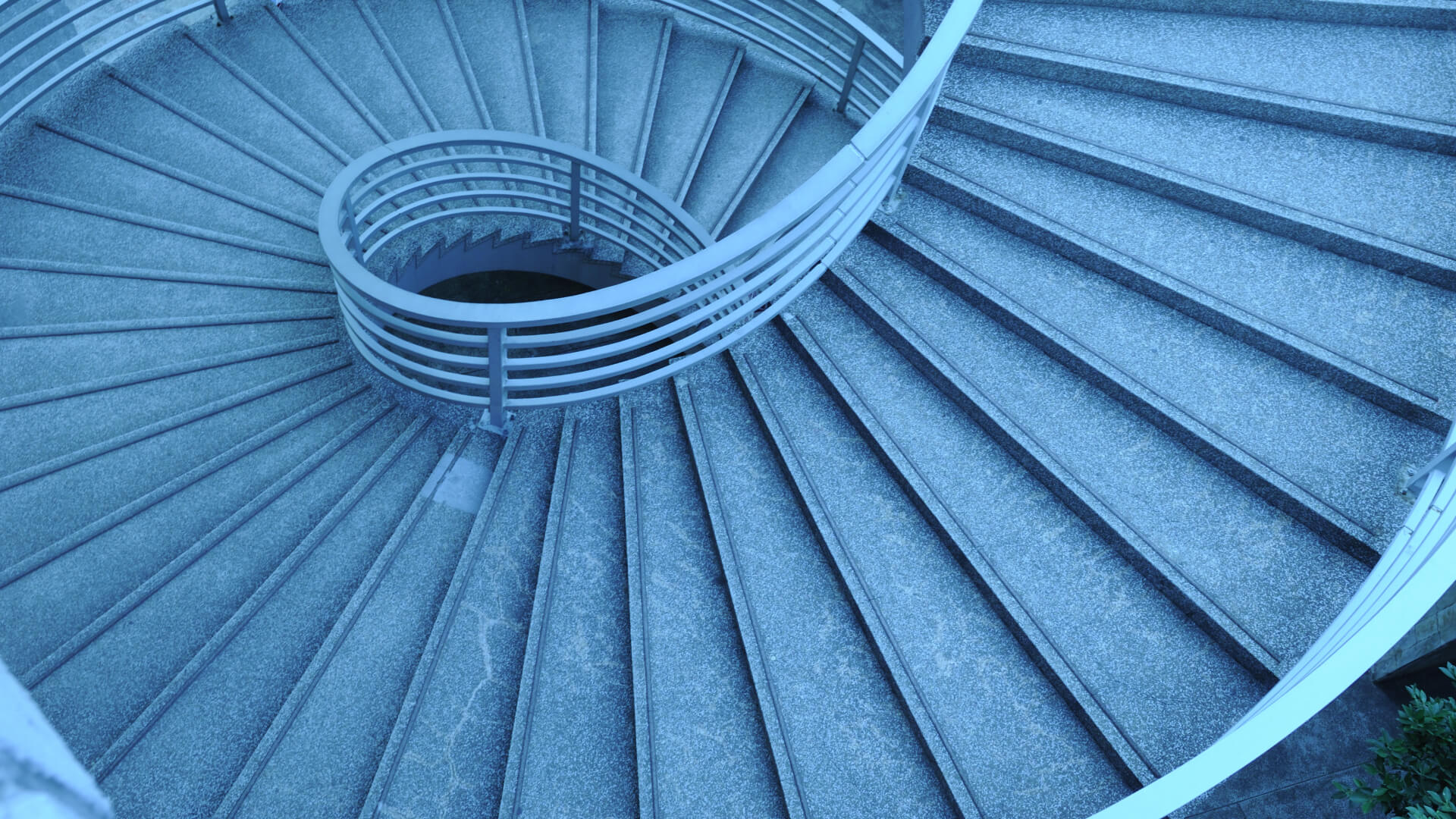 This image shows a striking spiral staircase with a sleek, modern design. The stairs are made of a metal material, likely steel or aluminum, with a distinctive spiral shape that draws the eye inward. The staircase has a cool, blue-toned color scheme that gives it a contemporary, minimalist aesthetic. The repetitive pattern of the spiral steps and the circular, open center create a visually captivating and almost hypnotic effect.