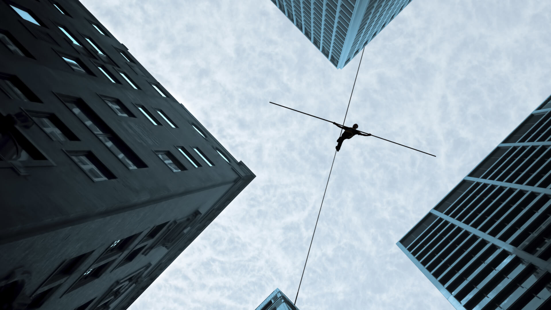 This image shows a dramatic upward view of a tightrope walker or high-wire artist balancing on a wire suspended between tall buildings in an urban setting. The perspective is from the ground looking up, with two skyscrapers framing the composition on either side against a cloudy sky. The silhouette of the person on the wire appears small against the vast scale of the buildings, creating a striking contrast. The image has a bluish-grey tonal quality that adds to its dramatic atmosphere.